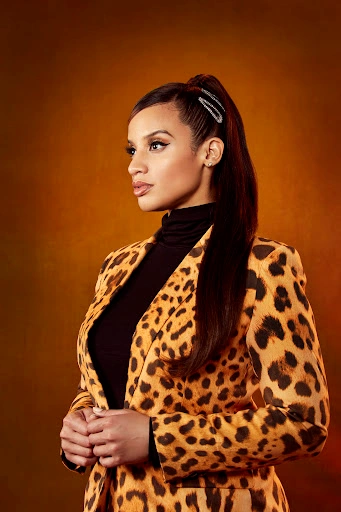 Woman wearing a leopard print blazer and black turtleneck with her hair in a high ponytail against an ochre textured backdrop