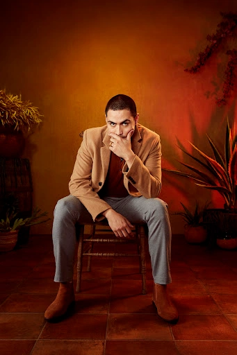 Man seated on a wooden chair wearing a camel coat, grey trousers, and brown boots against a warm-toned background