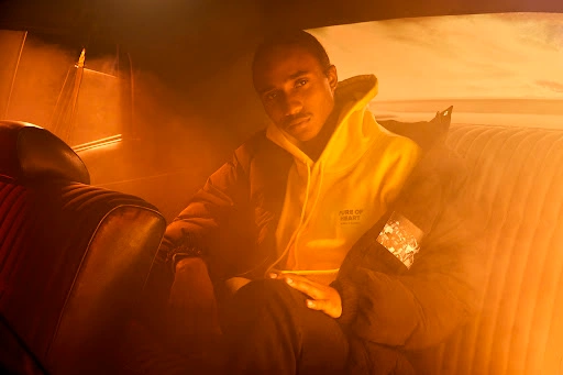A Black man seated inside the back of a car bathed in warm orange light, wearing a hoodie and a jacket.