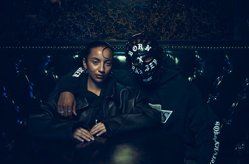 Two people seated together in a dark booth, a woman in a black leather jacket and a person wearing a patterned black hockey mask.