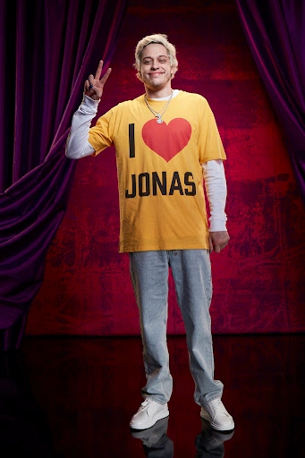 Pete Davidson wearing a yellow I Love Jonas t-shirt and light blue jeans flashing a peace sign in a portrait by Gizelle Hernandez.