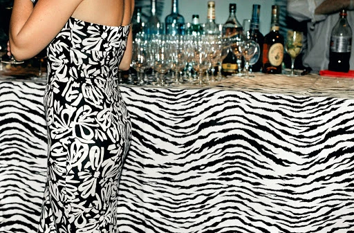 A woman in a strapless black and white dress stands in front of a bar covered in a zebra print tablecloth, glasses, and bottles.