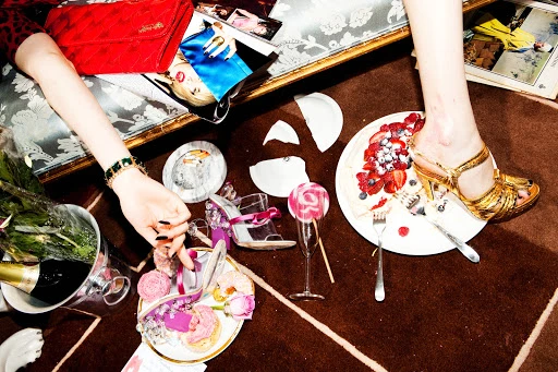Overhead shot of a woman's leg with a golden high heel stepping on a berry tart, surrounded by luxury accessories and broken dishes on a rug.