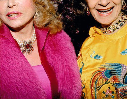 Two elegantly dressed older women pose with visible jewelry and bright, formal attire in a close-up shot, likely at an event.