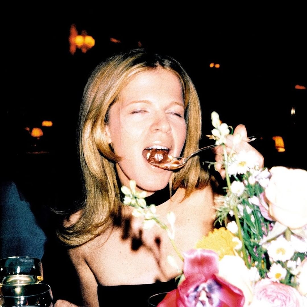 Blonde woman enjoys dessert with flowers at a dimly lit indoor event, captured in a candid snapshot with a spoon and a delighted expression.