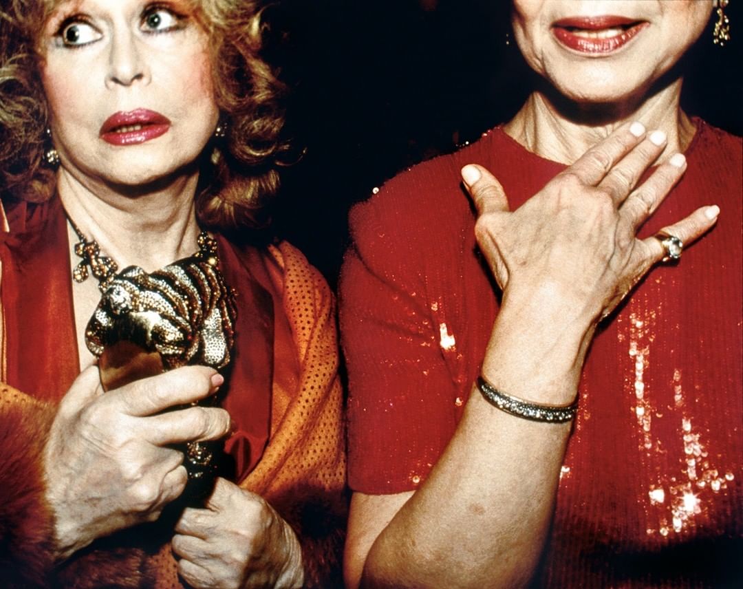 Two older women in red dresses at an event; one holds a dog-shaped purse. Dark backdrop, close shot, and rich textures highlight the scene.