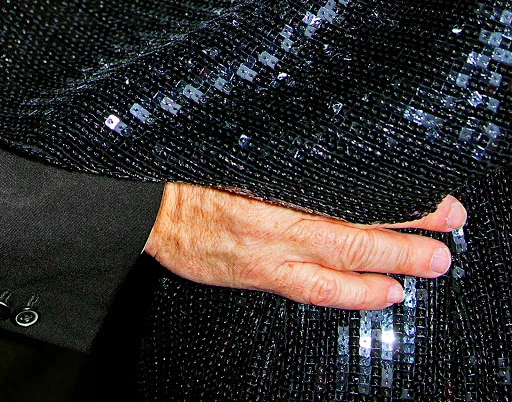 Closeup of a hand resting on a sequined jacket sleeve with a black suit jacket visible. The jacket has a shiny, textured appearance.
