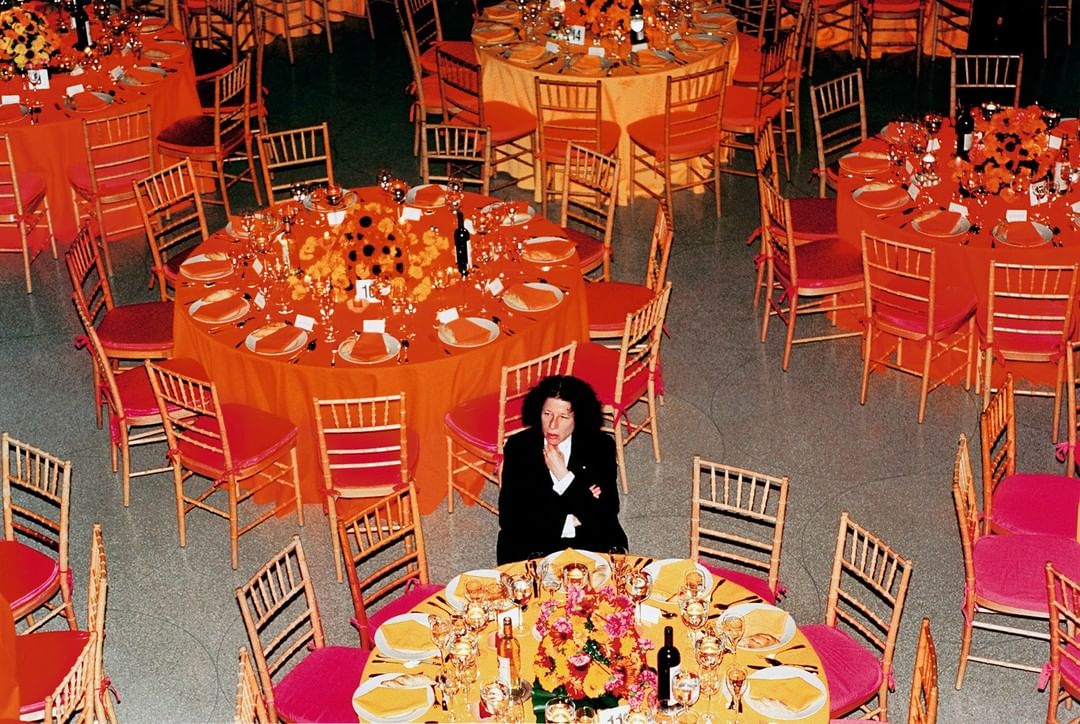 Overhead shot of an adult in a black suit seated at a formally set table in a banquet hall, surrounded by similar tables and chairs.