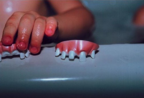 A child's hand holds dentures in a bathroom setting, creating a surreal and slightly unsettling yet intriguing visual composition.
