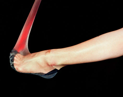 A foot and leg with hosiery is visible against a black background in a studio shot showcasing fashion and beauty.