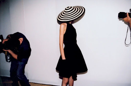 A woman in a black dress and a black and white striped hat poses against a white wall, with photographers on either side.