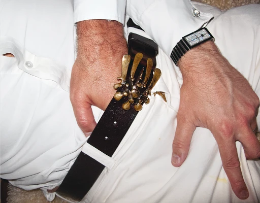Close-up of a man in a white shirt and pants, wearing a black leather belt with a gold ornamental buckle and a black wristwatch.