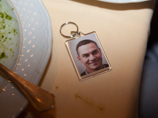 Close-up of a keychain with a portrait of a smiling adult male, sitting on a tan tablecloth next to a white plate and spoon.