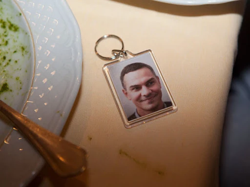 Close-up of a keychain with a portrait of a smiling adult male, sitting on a tan tablecloth next to a white plate and spoon.