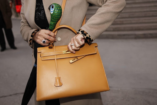 A tan leather Kelly bag with gold hardware is held by a woman wearing a beige coat. A decorative umbrella is in the background.