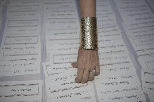 Close-up on a fair-skinned adult hand with a gold hammered cuff bracelet and ring, selecting from a stack of name cards.