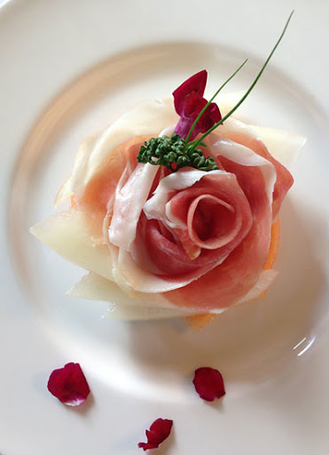 Close-up of an appetizer with melon, prosciutto rose, greens, and flower petals on a white plate, arranged for a sophisticated presentation.