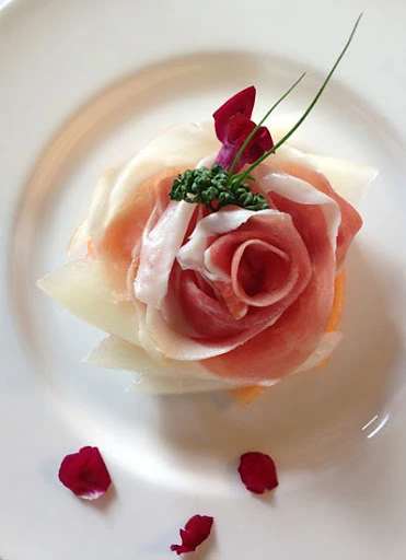 Close-up of an appetizer with melon, prosciutto rose, greens, and flower petals on a white plate, arranged for a sophisticated presentation.