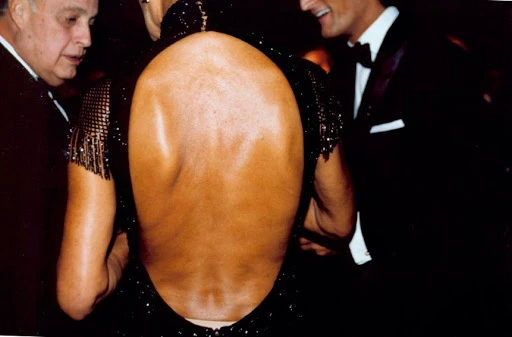 Back view of a woman in a black open-back dress with a beaded shoulder design, photographed at an indoor event with two men.