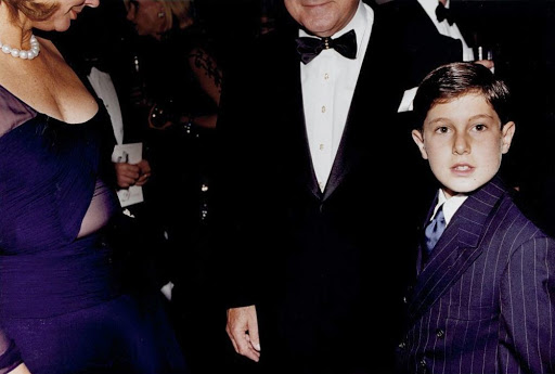 Formal gathering featuring a child in a pinstripe suit with adults in evening wear, captured in a dimly lit, indoor setting.