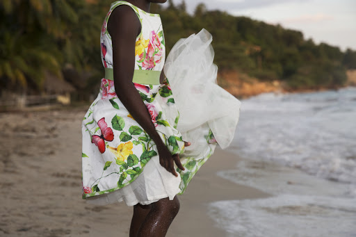Woman in floral dress on beach with white fabric blowing in wind. Ocean waves and tropical trees in the background at dusk.
