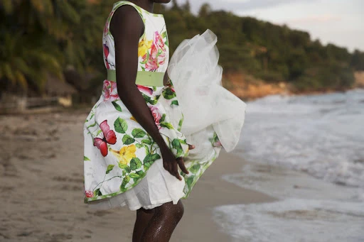 Woman in floral dress on beach with white fabric blowing in wind. Ocean waves and tropical trees in the background at dusk.