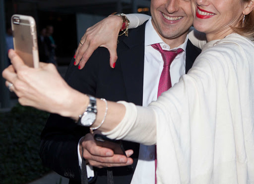 Smiling man and woman posing for a selfie in an outdoor setting. The woman holds a smartphone to capture the image.
