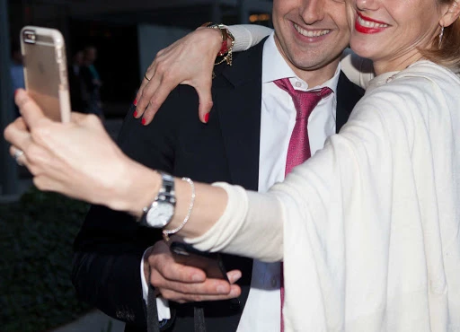 Smiling man and woman posing for a selfie in an outdoor setting. The woman holds a smartphone to capture the image.