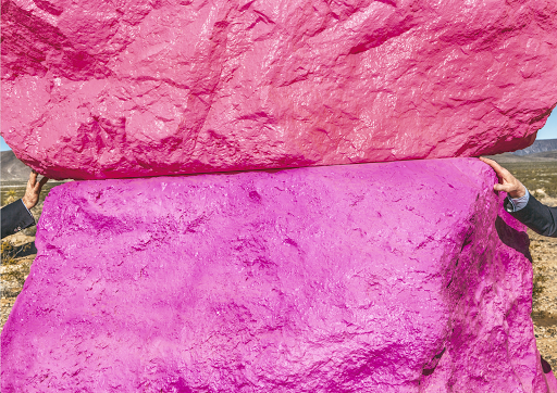 Two people hold up two large pink painted rocks outdoors in a desert landscape under a bright sky, creating a surreal, artistic scene.