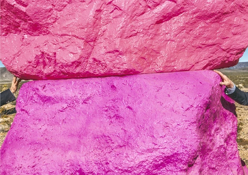 Two people hold up two large pink painted rocks outdoors in a desert landscape under a bright sky, creating a surreal, artistic scene.