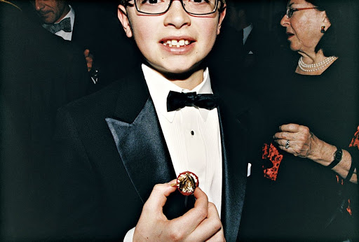 Young boy in a tuxedo adjusts a lapel pin at an event, surrounded by adults in formal wear, captured in a medium shot.