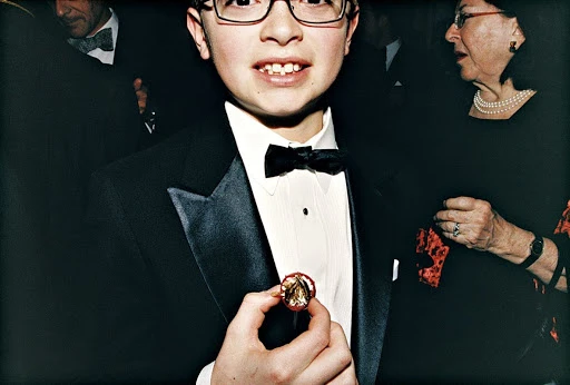 Young boy in a tuxedo adjusts a lapel pin at an event, surrounded by adults in formal wear, captured in a medium shot.