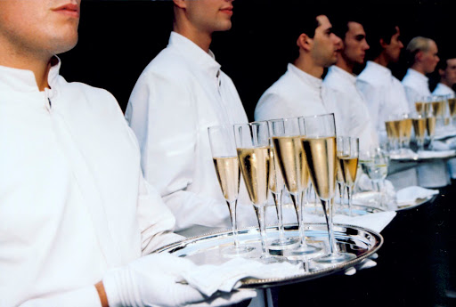 Row of servers in white jackets hold trays of champagne flutes. The champagne is golden. Elegant and formal setting.