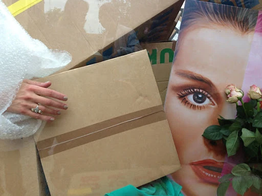 Woman holding brown packing box near a face poster with roses, bubble wrap, and U-Haul box in indoor setting, natural lighting.