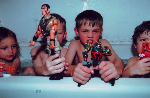 Four children in a bathtub filled with water, holding action figure toys, with a bright, direct, and unfiltered style.