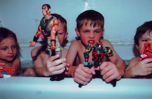 Four children in a bathtub filled with water, holding action figure toys, with a bright, direct, and unfiltered style.