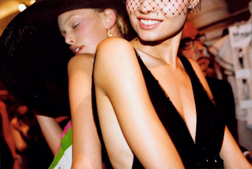 Two women pose intimately, one in a black dress with a veil, the other in a black hat with a colorful top, back stage at a fashion event.