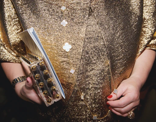 A woman in a shiny gold outfit holds a translucent cosmetic case and gold necklace in an indoor setting, displaying a glamorous aesthetic.