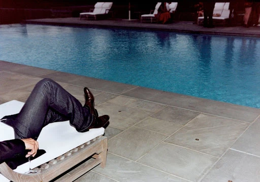 Man lounges poolside on a white chair with sunglasses and a dark jacket, overlooking a large swimming pool surrounded by lounge chairs.