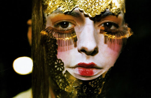 Artistic close-up of a face with gold headpiece, dramatic eyelashes, and red lips against a dark background. Beauty and makeup concept.