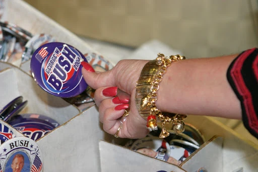 Hand holding a George W. Bush campaign button from 2004, displayed in a cardboard box, accessorized with gold bracelet and red manicure.