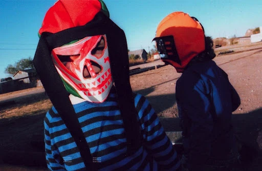 Two children wearing colorful masks stand outdoors on a sunny day. One mask is red and white, the other is orange and black.