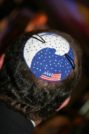 Person wearing a blue and white kippah with an American flag design on the back of their head at an indoor event.