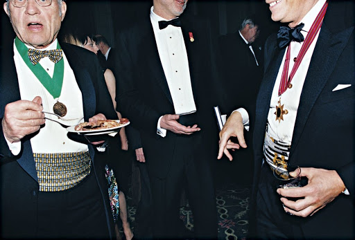 Three formally dressed men stand in a group setting at an indoor event. One man holds a plate of food, wearing a green sash and medal.
