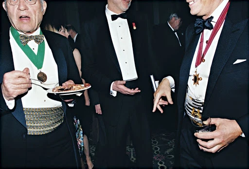 Three formally dressed men stand in a group setting at an indoor event. One man holds a plate of food, wearing a green sash and medal.