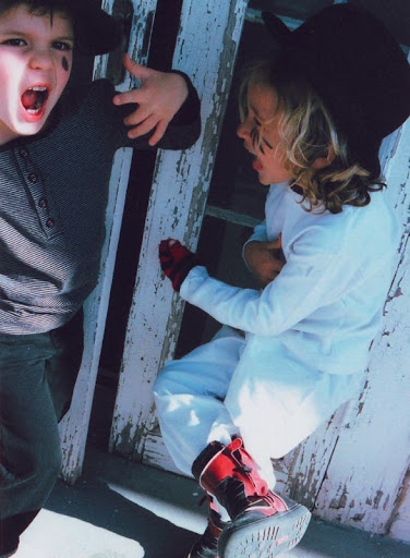 Two young children in hats with face paint pretend fight in front of weathered white wood, one with open mouth and red gloves. 