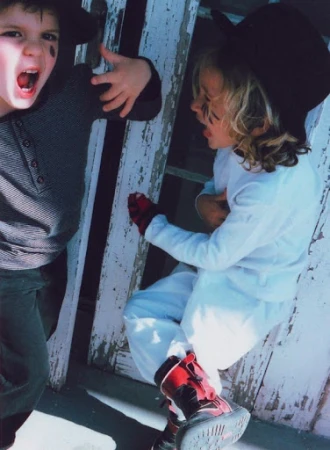 Two young children in hats with face paint pretend fight in front of weathered white wood, one with open mouth and red gloves. 