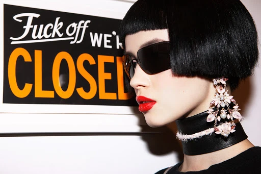 Profile shot of a model with blunt black bob, sunglasses, and statement earrings next to a sign that reads 'Fuck off we're CLOSED'.
