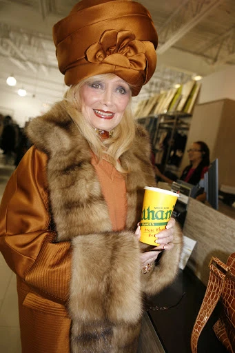 Elegant woman with blond hair and a large brown hat, wearing a fur-trimmed coat, holding a yellow cup with green logo indoors.