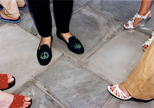 Close-up captures a diverse group of people's feet wearing an assortment of shoes and sandals on a stone-tiled surface.